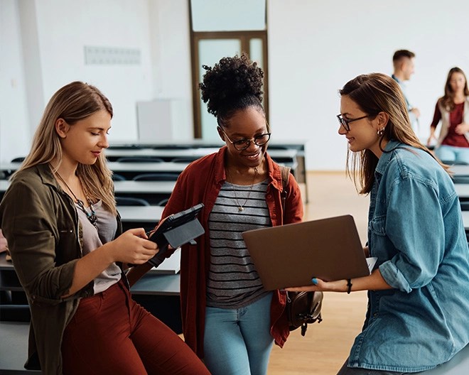 Three young university students in a classroom discussing content on a laptop and a tablet.