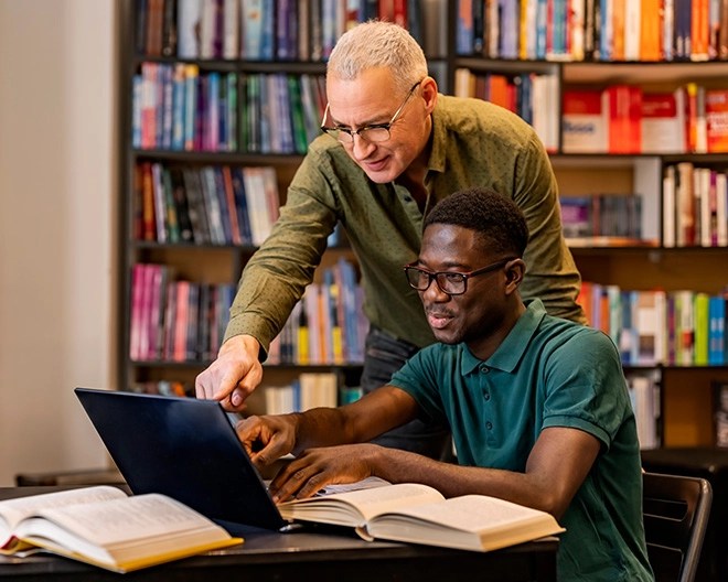 Teacher assists high school student in a library; the teacher points at a laptop while the student refers to open textbooks.