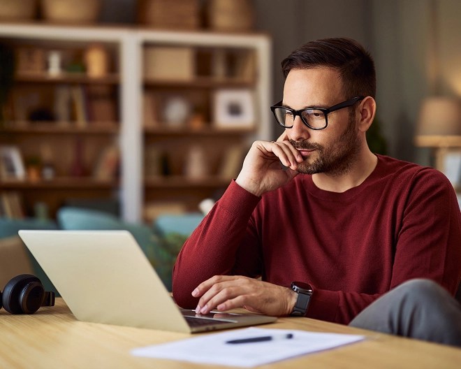 Online student wearing glasses and a red sweater, working on his laptop at home with his feet up.