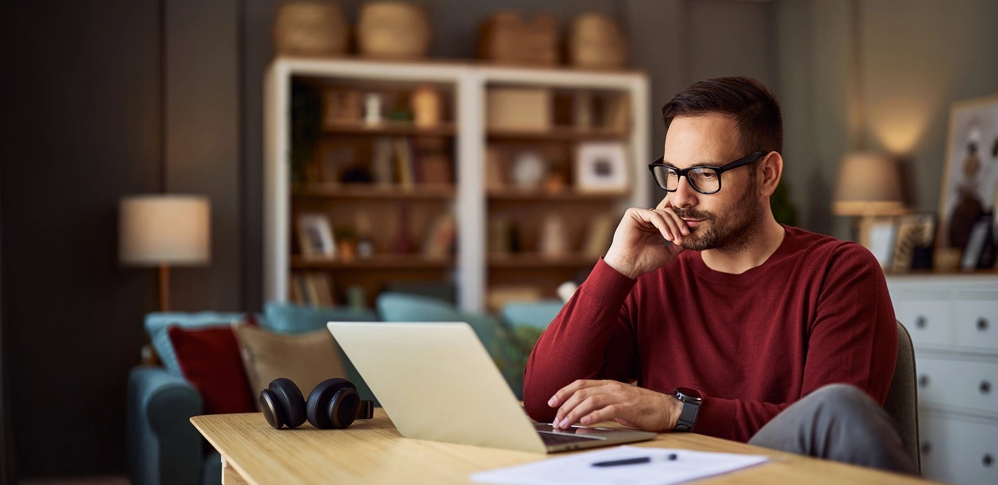 Online student wearing glasses and a red sweater, working on his laptop at home with his feet up.