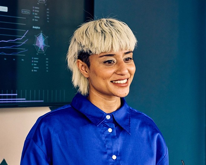 Young, smiling business professional wearing a bright blue collared top, standing in front of a screen displaying data figures.