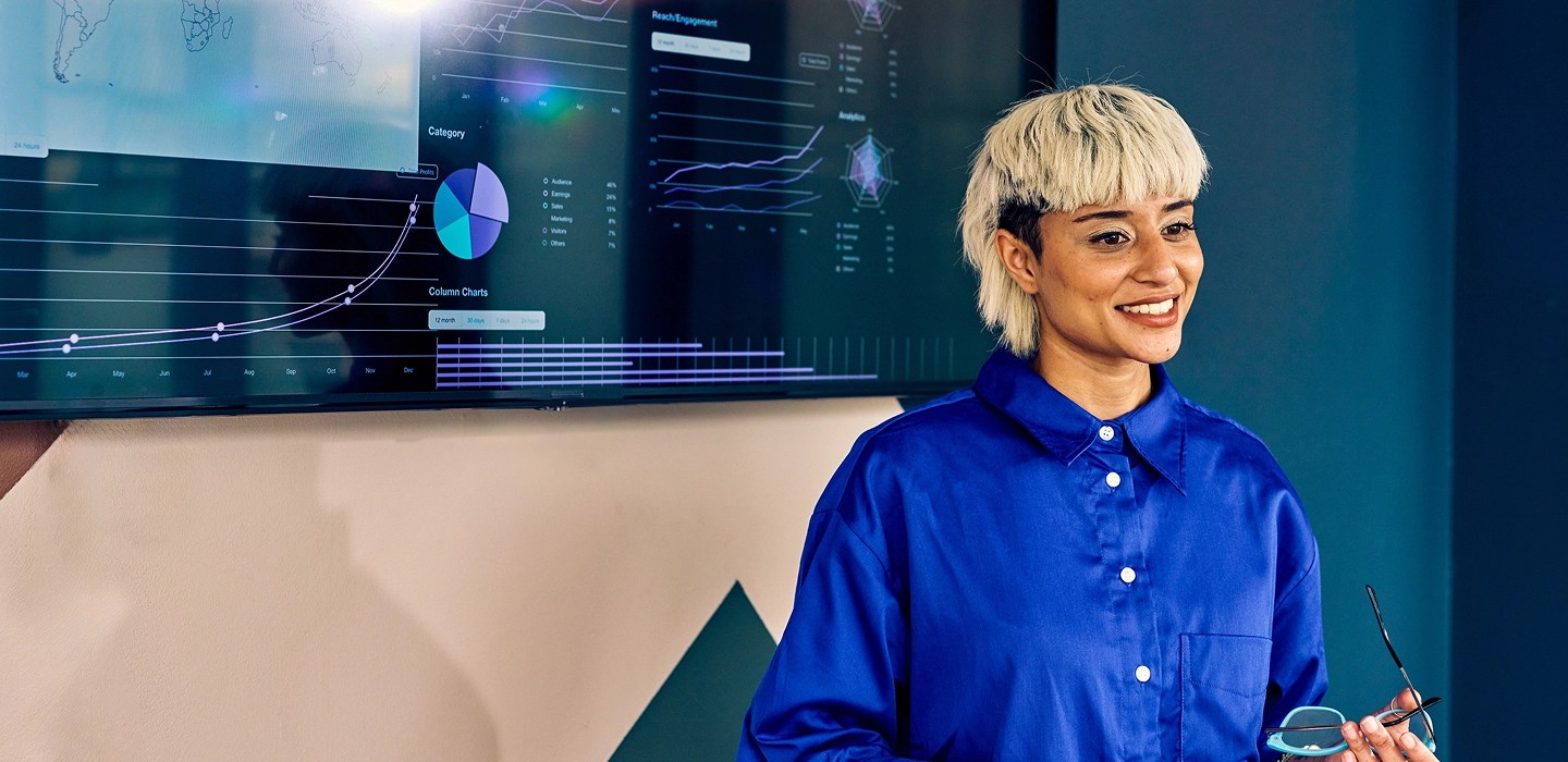 “A woman with short blonde hair wearing a blue button-up shirt, standing in front of a large screen displaying graphs.