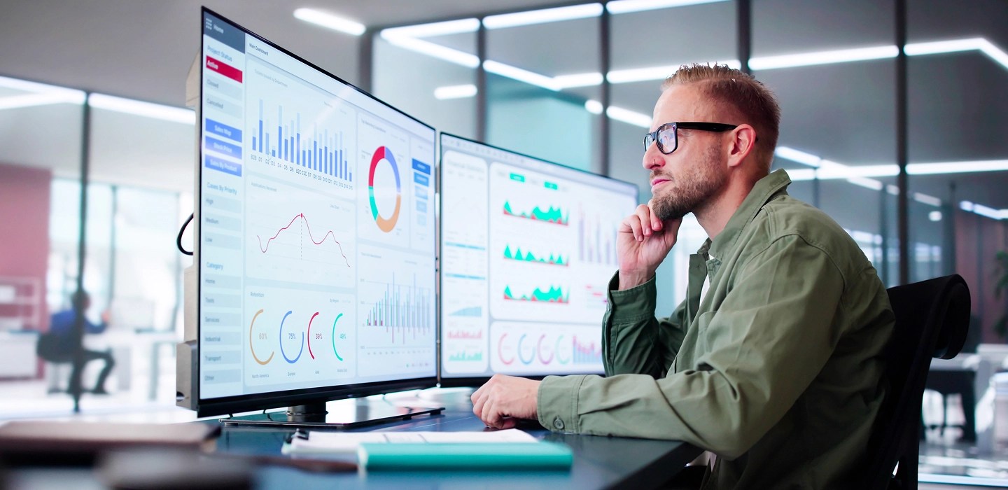 Financial professional seated at a desk, analysing economic charts and figures displayed on dual monitors in a well-lit office.