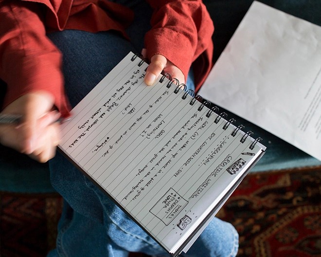 Close-up of a person writing in a notebook filled with handwritten text and doodles.