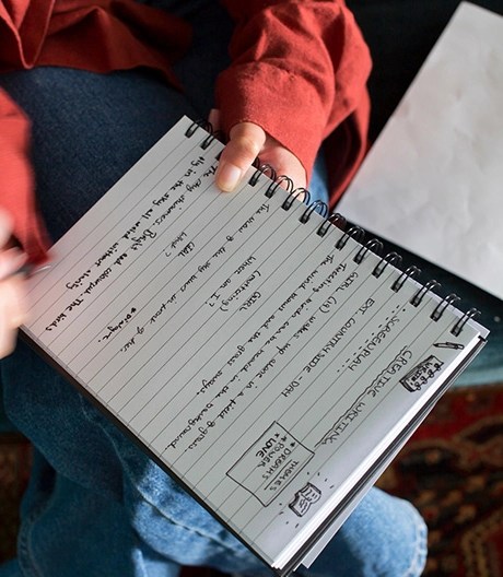 Close-up of a person writing in a notebook filled with handwritten text and doodles.
