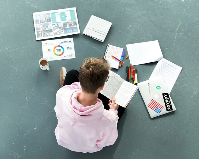 Bird’s-eye view of a marketing student reading a notebook, surrounded by a laptop, markers, pencils, charts, and documents spread out on the floor.