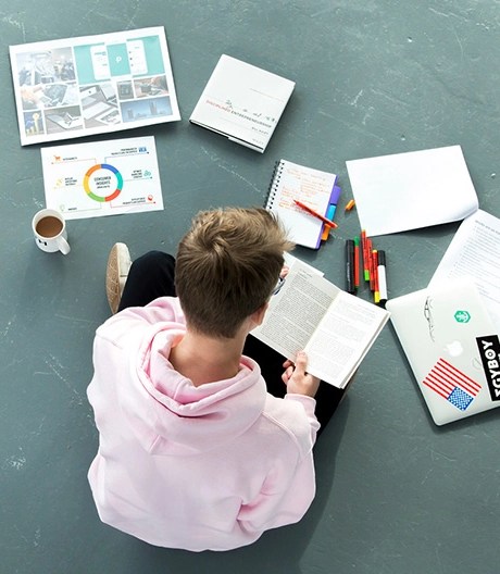 Bird’s-eye view of a marketing student reading a notebook, surrounded by a laptop, markers, pencils, charts, and documents spread out on the floor.