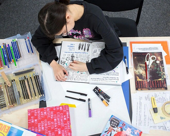 Bird’s-eye view of an art student looking down and illustrating in her notebook filled with sketches, surrounded by a desk full of art supplies.
