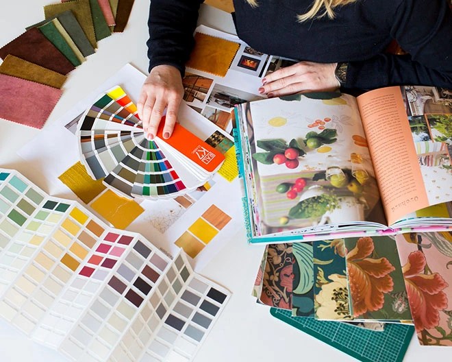 Close-up of an interior designer’s hands reviewing colour palettes, fabric swatches, wallpaper samples, and an open notebook with design inspiration.