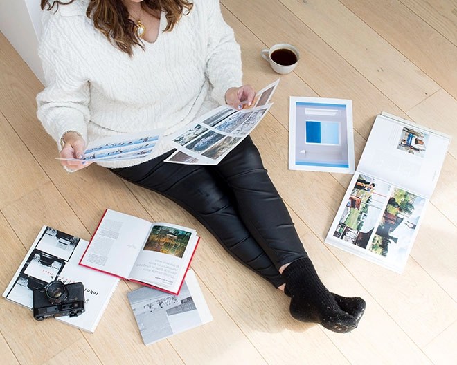 Woman seated on wooden floor in a white cable‑knit sweater reviewing photo prints, surrounded by open photobooks, a vintage camera and a cup of coffee.