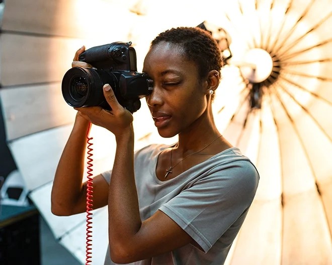 A young photographer in a grey short-sleeved t-shirt captures a photo with focused intent, a large studio light glowing softly in the background.
