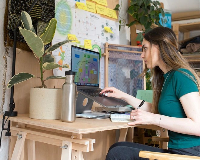 Fashion student taking notes on sustainable fashion while watching a presentation on her computer, with a large bulletin board covered in charts and Post-It notes behind her desk.