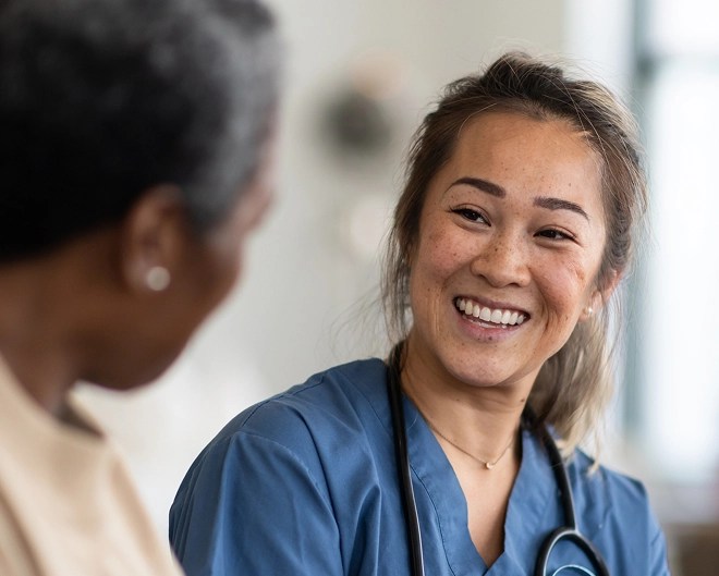 Friendly doctor in scrubs with a stethoscope around her neck, smiling warmly at a patient seated in the foreground, in a bright clinical setting.
