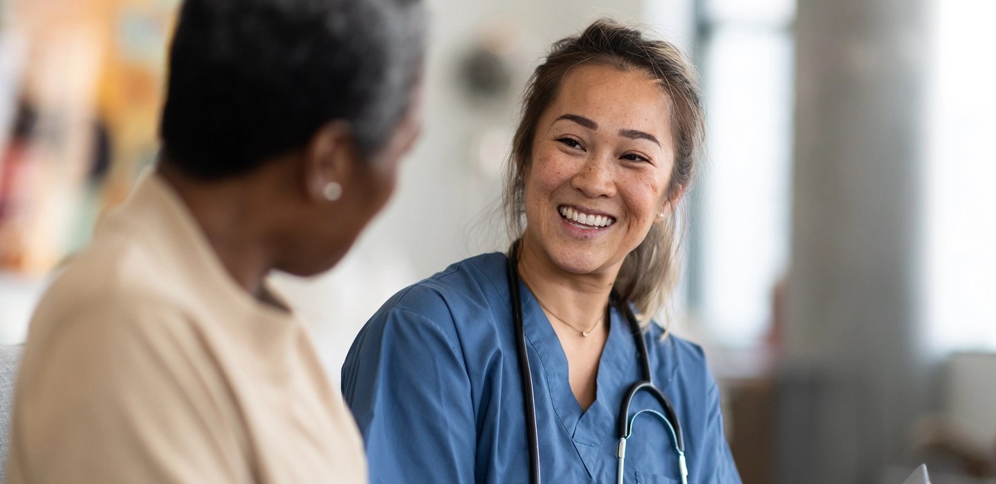 Friendly doctor in scrubs with a stethoscope around her neck, smiling warmly at a patient seated in the foreground, in a bright clinical setting.