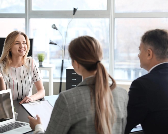 Two managers—one in a suit and the other in a plaid blazer holding a clipboard—speak with a direct report wearing a striped top in a bright office.