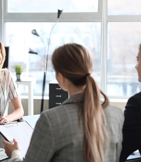 Two managers—one in a suit and the other in a plaid blazer holding a clipboard—speak with a direct report wearing a striped top in a bright office.