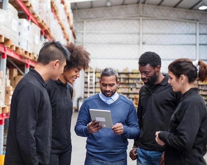 Facilities manager discusses a topic on a tablet with four young floor workers in a bright warehouse.