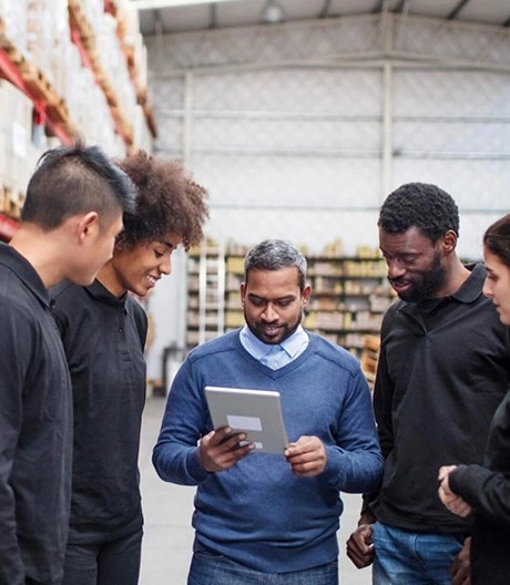 Facilities manager discusses a topic on a tablet with four young floor workers in a bright warehouse.