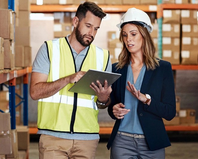 A floor worker wearing a neon safety vest and holding a clipboard discusses with a floor manager in a blazer and hard hat inside a warehouse.