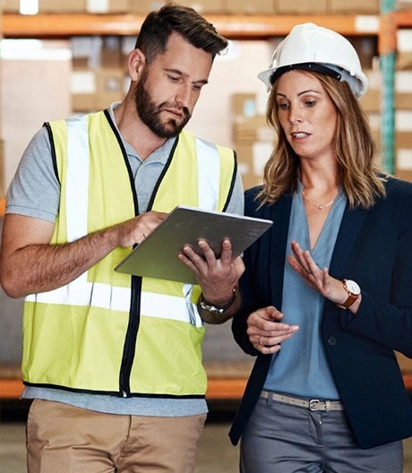 A floor worker wearing a neon safety vest and holding a clipboard discusses with a floor manager in a blazer and hard hat inside a warehouse.