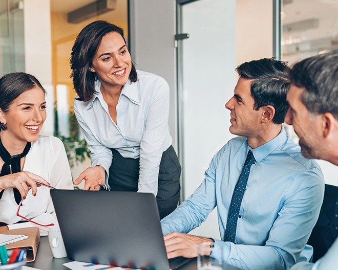 Four coworkers in business casual clothing smile and engage in discussion during a meeting in a bright office.