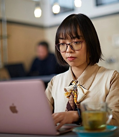 Online student with a collared blouse and glasses works on her laptop in a bright cafe.