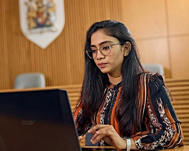 Young professional with long hair, glasses, and a patterned top, working on her laptop inside a courtroom.