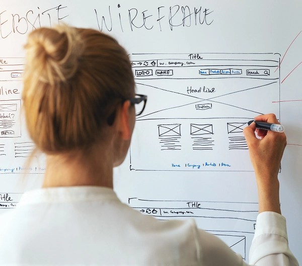 Close-up of a woman with her hair up, wearing a white blouse, sketching a wireframe layout on a whiteboard.