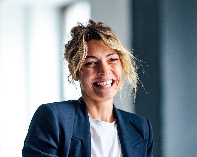 Close-up of a smiling businesswoman wearing a navy blue blazer and white t-shirt, holding a clipboard in an office setting.