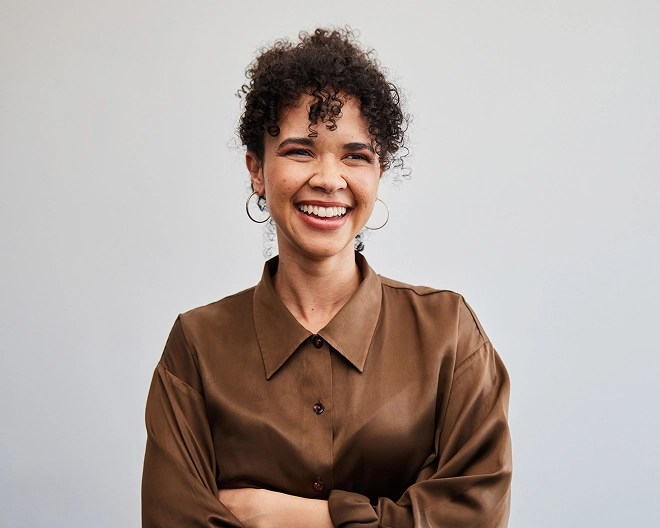 Close-up headshot of a young professional with curly hair, wearing a brown collared top and earrings, smiling with her arms crossed.