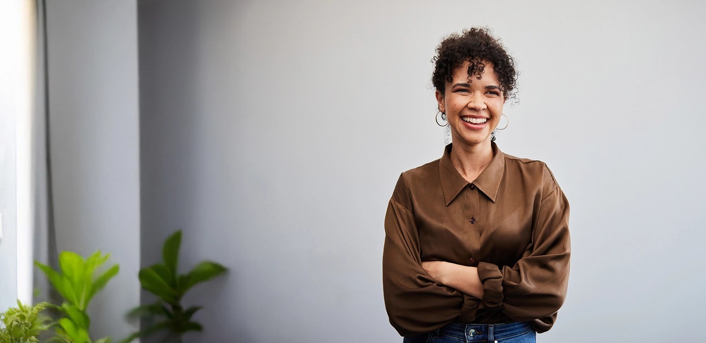 Close-up of a young professional with curly hair, wearing a brown collared top and earrings, smiling with her arms crossed.