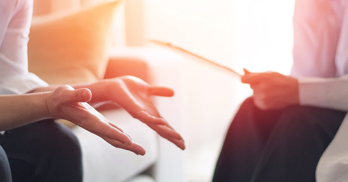 Close-up of hands gesturing during a counselling session, with a therapist holding a notepad in the background.