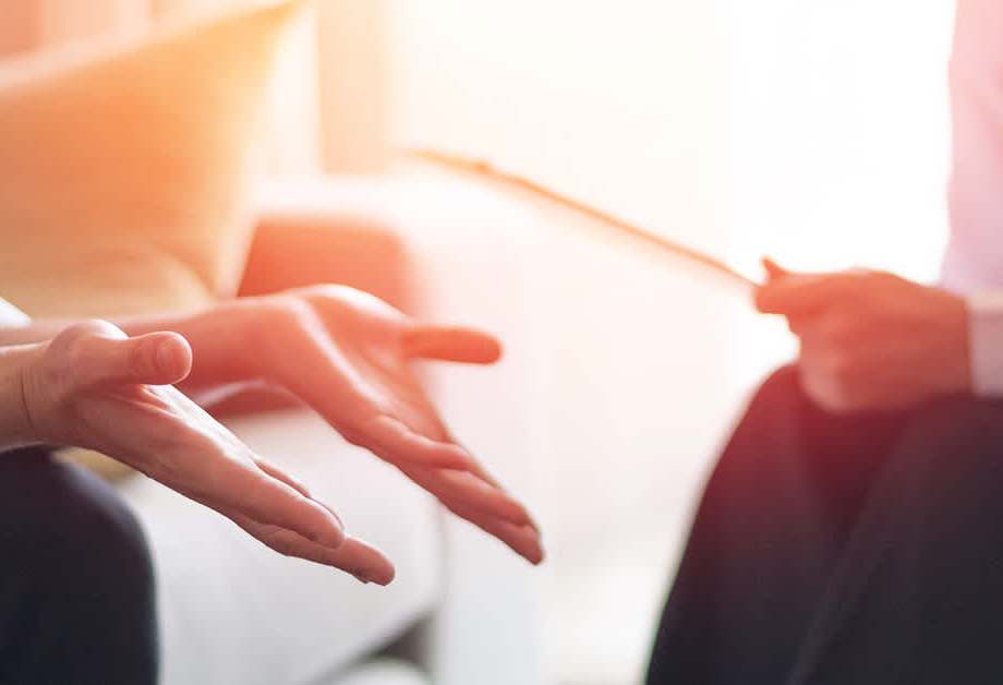 Close-up of hands gesturing during a counselling session, with a therapist holding a notepad in the background.