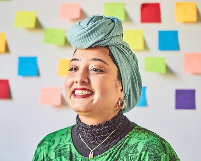 Close-up of a smiling marketing professional wearing a sea green headwrap and matching top, standing in front of a whiteboard covered in multi-coloured sticky notes.