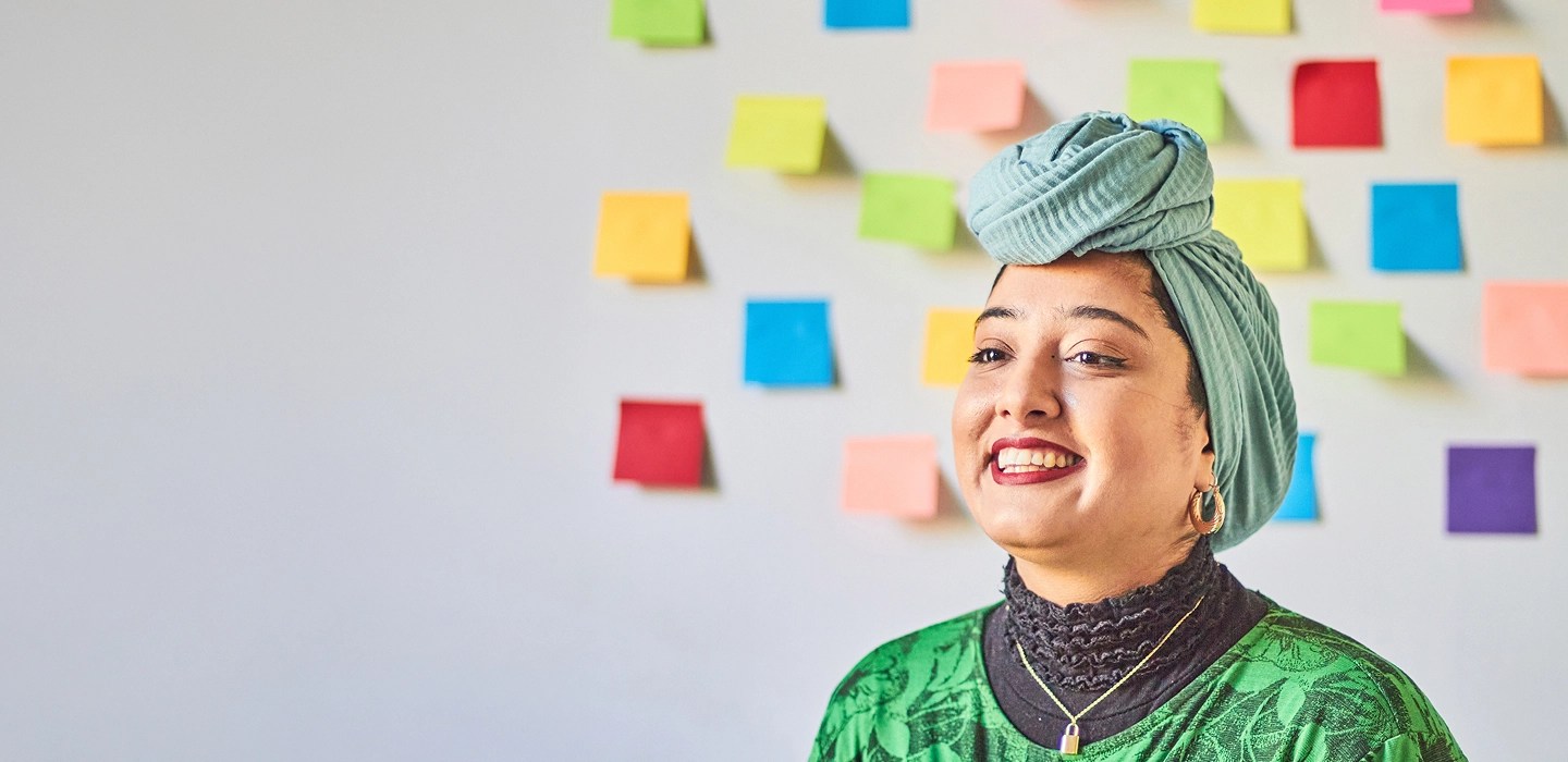 A woman in a green hijab smiling, with sticky notes on the wall behind her.