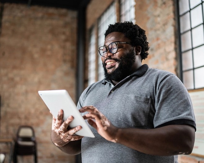 Cheerful professional standing and working on a tablet in a bright room with exposed brick walls.