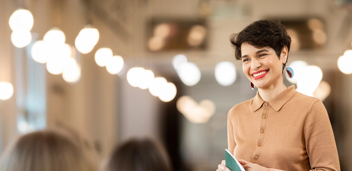 Smiling young professional with short hair, wearing a tan collared sweater and bright lipstick, holding a green notebook.