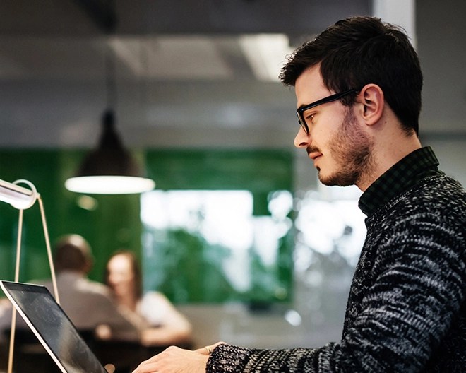 Young professional wearing glasses and a sweater, focused on his laptop while working in a modern co-working space.