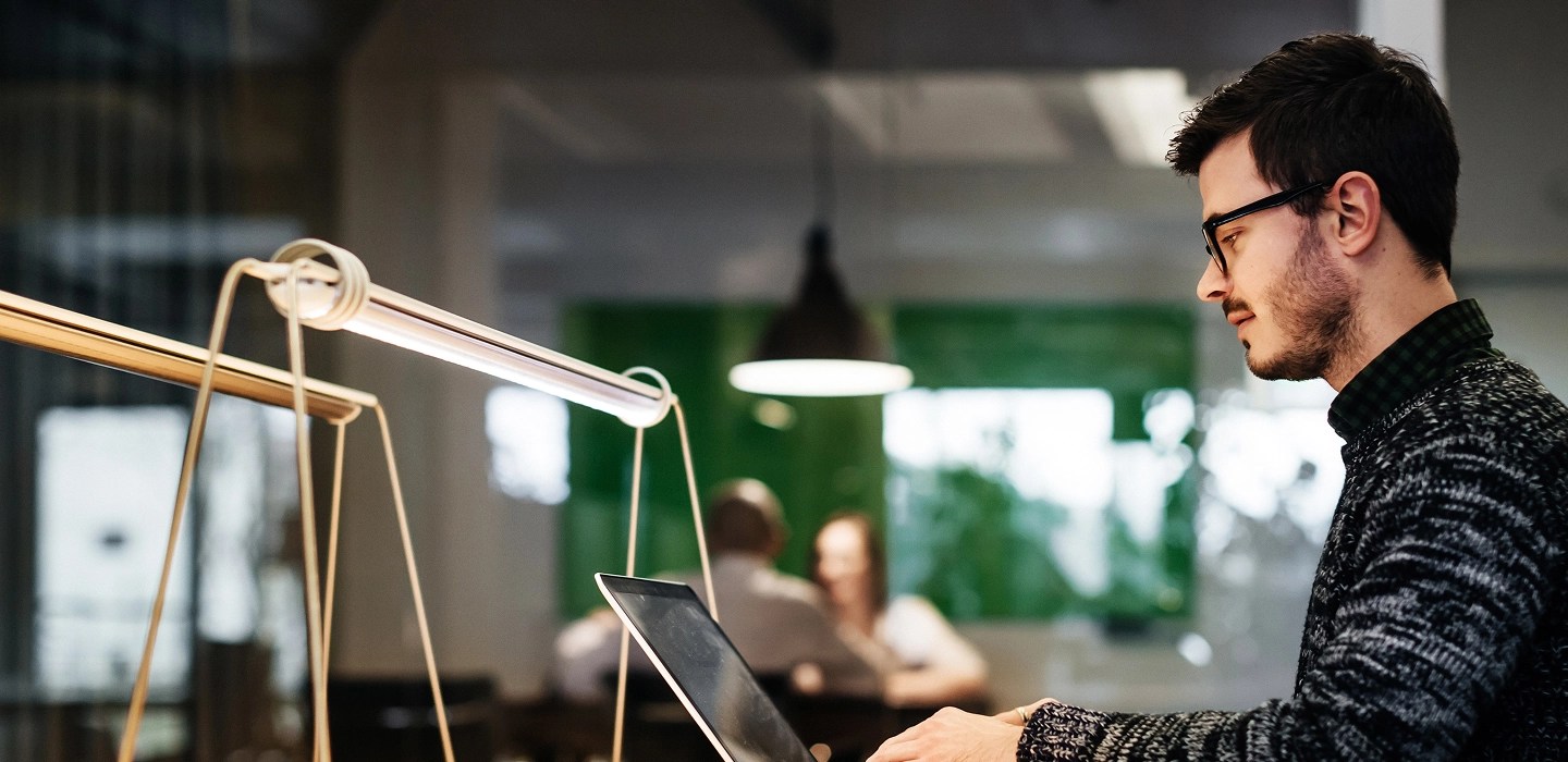 Young professional wearing glasses and a sweater, focused on his laptop while working in a modern co-working space.