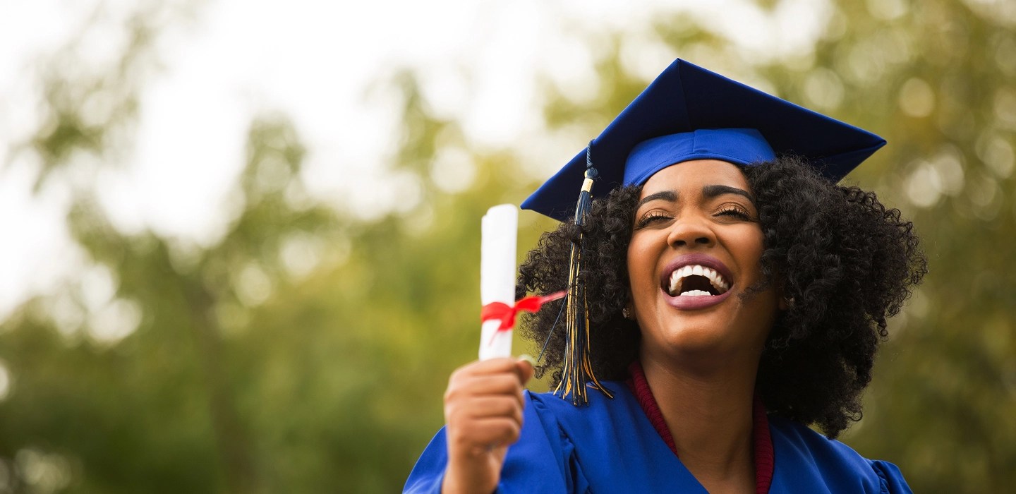 Elated and smiling graduate wearing a blue graduation cap and gown holds her diploma proudly.