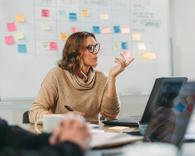Professional woman in a cowl-neck sweater and glasses holds a pen and gestures during a meeting in a bright office. The table is covered with notebooks, papers, and open laptops, with a whiteboard full of sticky notes blurred in the background.