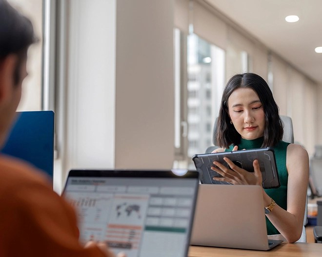 Two colleagues work independently in a shared office space; one in the foreground is slightly out of frame and blurred while analysing charts and maps on his open laptop, while the other, wearing a green tank top with short brown hair, is more in focus and works on her tablet.