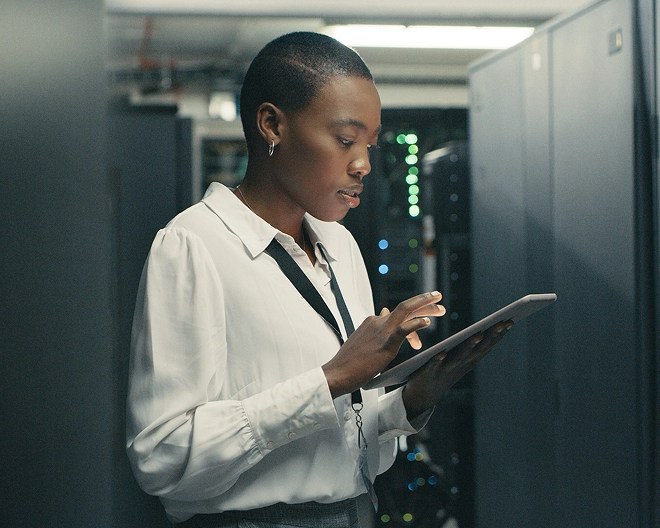 IT professional analyses data on a tablet while standing in a server room surrounded by equipment and blinking lights.