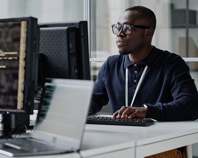 IT professional wearing glasses and a collared shirt focuses on his computer screen at a shared desk in a modern office setting.