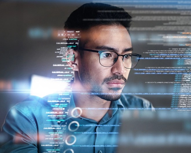 Close-up of an IT professional wearing glasses, intently viewing his computer screen, with a digital overlay of code and data reflected around his face.
