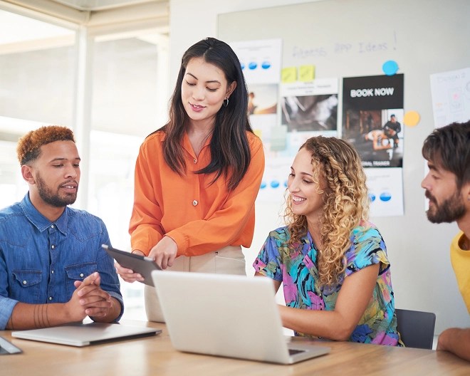 Marketing professional with long brown hair and a bright orange blouse enthusiastically presents something on her tablet to her smiling team in a bright office setting.