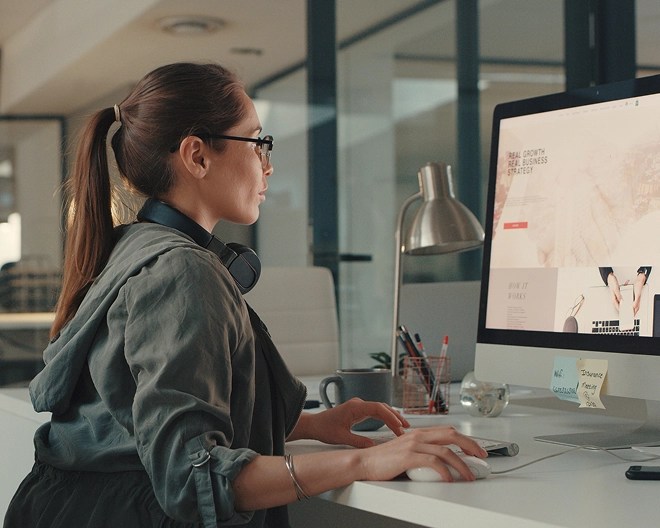 Young professional in a modern office, dressed in casual clothes with headphones around her neck, focuses on designing or editing a website on her desktop computer.