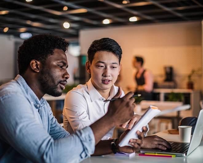 Two male colleagues collaborate in a shared office space. The one in the foreground holds a notebook and pencil, focused on his notes, while the other listens attentively to his coworker’s ideas.