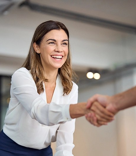 Close-up of a young businesswoman with shoulder-length dirty blonde hair wearing a white blouse, smiling warmly as she shakes hands with someone just out of frame, suggesting a successful meeting or introduction.