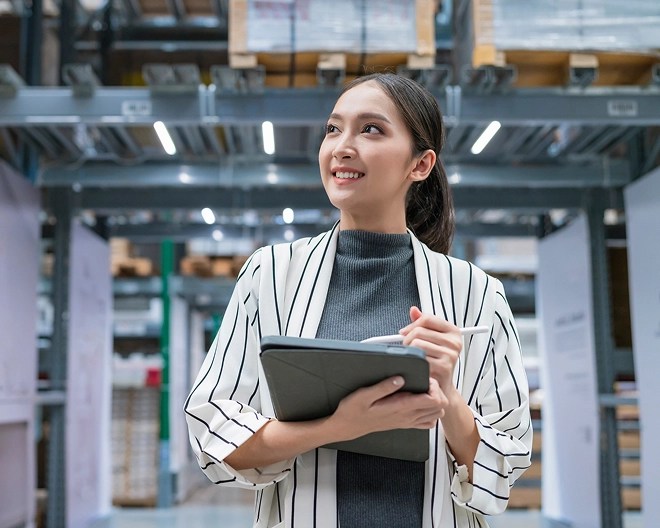 Young professional in a white and grey striped cardigan and grey top, her long brown ponytail pulled back neatly, smiles as she looks up and off to something out of frame. She holds a tablet and stylus confidently in a bright warehouse setting, suggesting active engagement or collaboration.